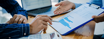 Two people pointing to a clipboard holding a report with charts.