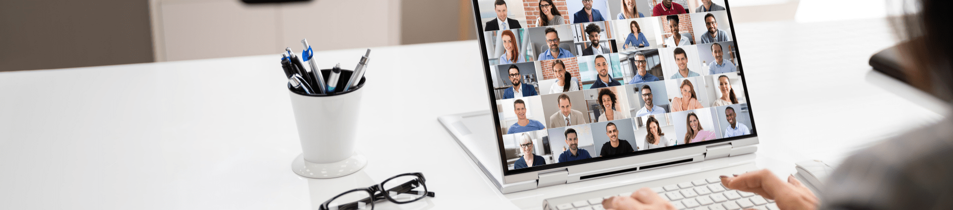 An over-the-shoulder view of someone's desk with a cup of pens, a pair of reading glasses, and a laptop featuring an online meeting.